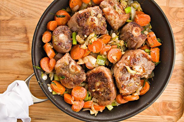 "An overhead close up of a large frying pan containing a colorful mixture of browned oxtails and vegetables, the makings for a flavorful  oxtail soup. Isolated on a wooden surface."